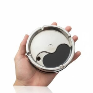 Close-up of a small, round, stainless steel metal ashtray with a rotating lid featuring a black and silver Yin and Yang design, held in the palm of a hand against a white background.
